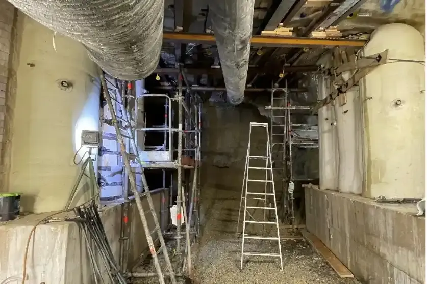 Das Bild zeigt den Blick in einen Abbau im Bergwerk. Auf der linken und rechten Seite stehen jeweils drei helle, säulenartige Stützbauwerke auf einem grauen Betonfundament. Quer über die Stützbauwerke, direkt unterhalb der Decke, verlaufen dunkle Metallträger. Zwischen den Stützbauwerken stehen Leitern und Gerüste. Im Hintergrund und auf dem Boden liegt Schotter.