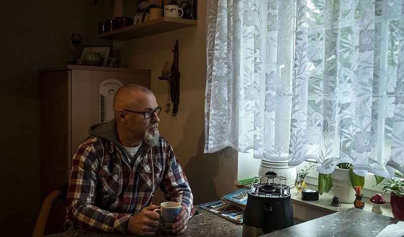 Foto eines Mannes mit kurzen Haaren und grauem Bart, der an einem Küchentisch mit einer Tasse in den Händen sitzt. Der Mann trägt eine karierte Kapuzenjacke. Er schaut seitlich aus dem Fenster, vor dem weiße Gardinen hängen.