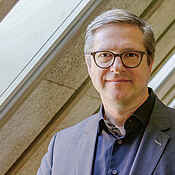 Portraitfoto eines Mannes mit dunkelblonden kurzen Haaren und Brille. Der Mann trägt ein dunkles Hemd und Sakko. Im Hintergrund sind unscharf Fenster und Dachschrägen zu sehen. 
