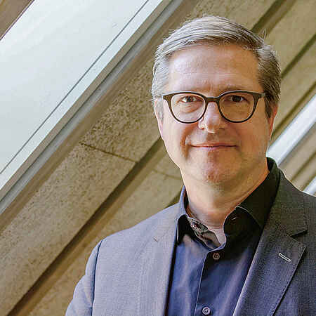Portraitfoto eines Mannes mit dunkelblonden kurzen Haaren und Brille. Der Mann trägt ein dunkles Hemd und Sakko. Im Hintergrund sind unscharf Fenster und Dachschrägen zu sehen. 