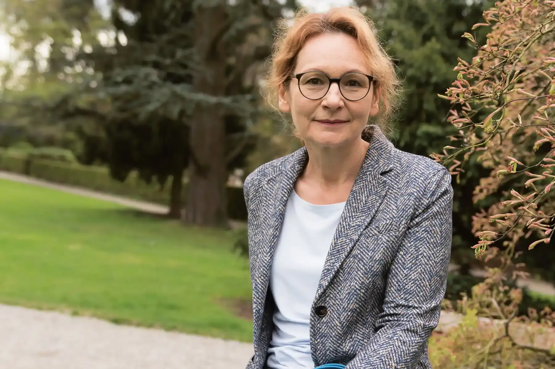 Portrait einer Frau mit Brille und roten Haaren, die aus dem Gesicht gebunden wurden. Sie trägt einen blau-weiß gemusterten Blazer über einem hellgrauen Oberteil. Im Hintergrund ist ein Park mit Grünflächen, Bäumen und Wegen zu sehen.