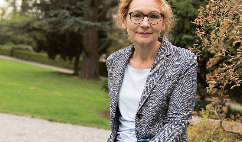 Portrait einer Frau mit Brille und roten Haaren, die aus dem Gesicht gebunden wurden. Sie trägt einen blau-weiß gemusterten Blazer über einem hellgrauen Oberteil. Im Hintergrund ist ein Park mit Grünflächen, Bäumen und Wegen zu sehen.