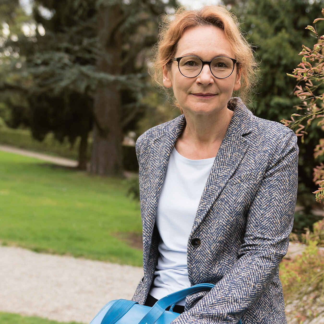 Portrait einer Frau mit Brille und roten Haaren, die aus dem Gesicht gebunden wurden. Sie trägt einen blau-weiß gemusterten Blazer über einem hellgrauen Oberteil. Im Hintergrund ist ein Park mit Grünflächen, Bäumen und Wegen zu sehen.