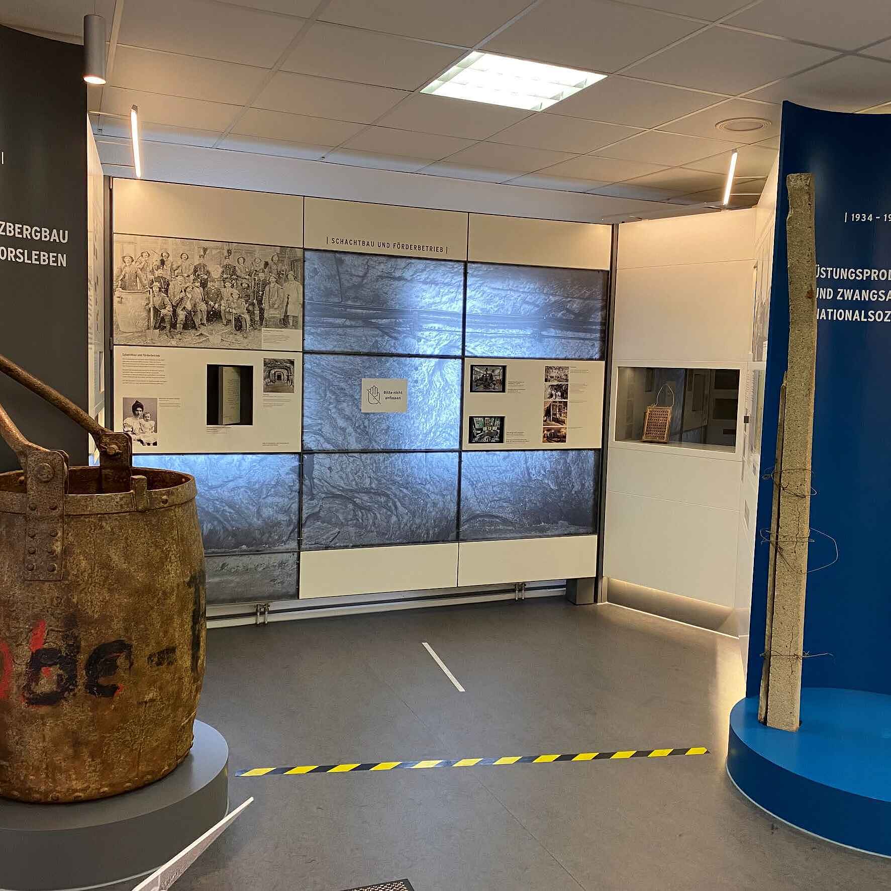 In einem Ausstellungsraum steht links im Bild ein Teufkübel, der aussieht wie ein sehr großer Metalleimer.