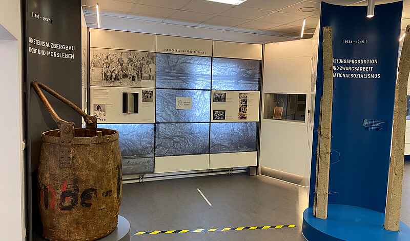 In einem Ausstellungsraum steht links im Bild ein Teufkübel, der aussieht wie ein sehr großer Metalleimer.