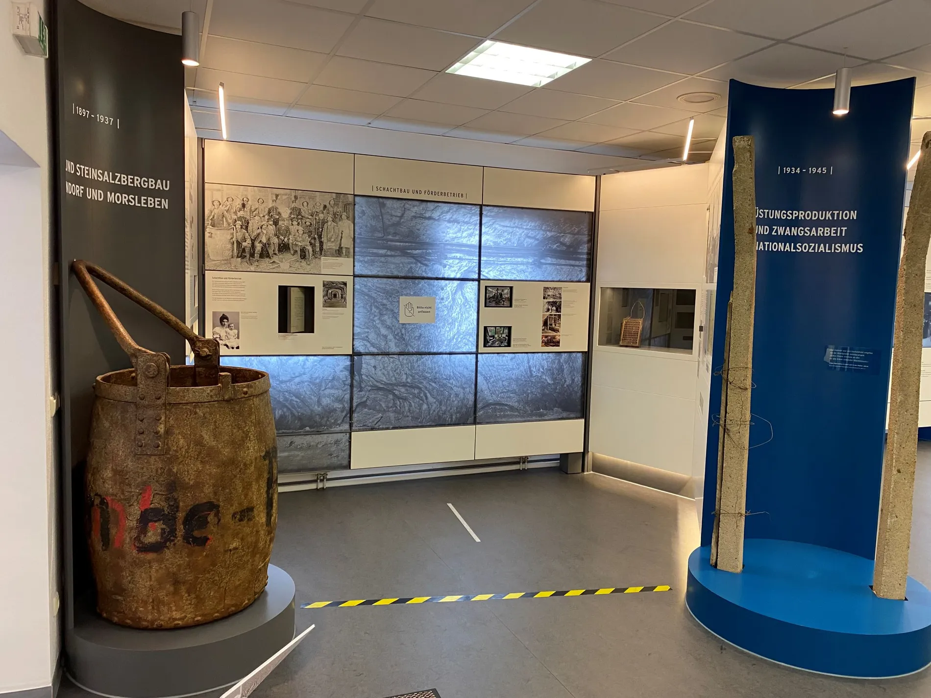 In einem Ausstellungsraum steht links im Bild ein Teufkübel, der aussieht wie ein sehr großer Metalleimer.