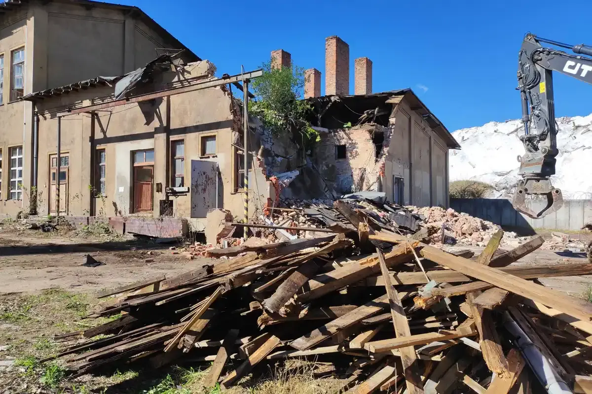 Foto eines Haufens alter, zersplitterter Holzbalken. Sie stammen aus einem verwitterten Gebäude, dessen Abriss begonnen hat. Links im Bild befindet sich der Arm des Abrissbaggers.
