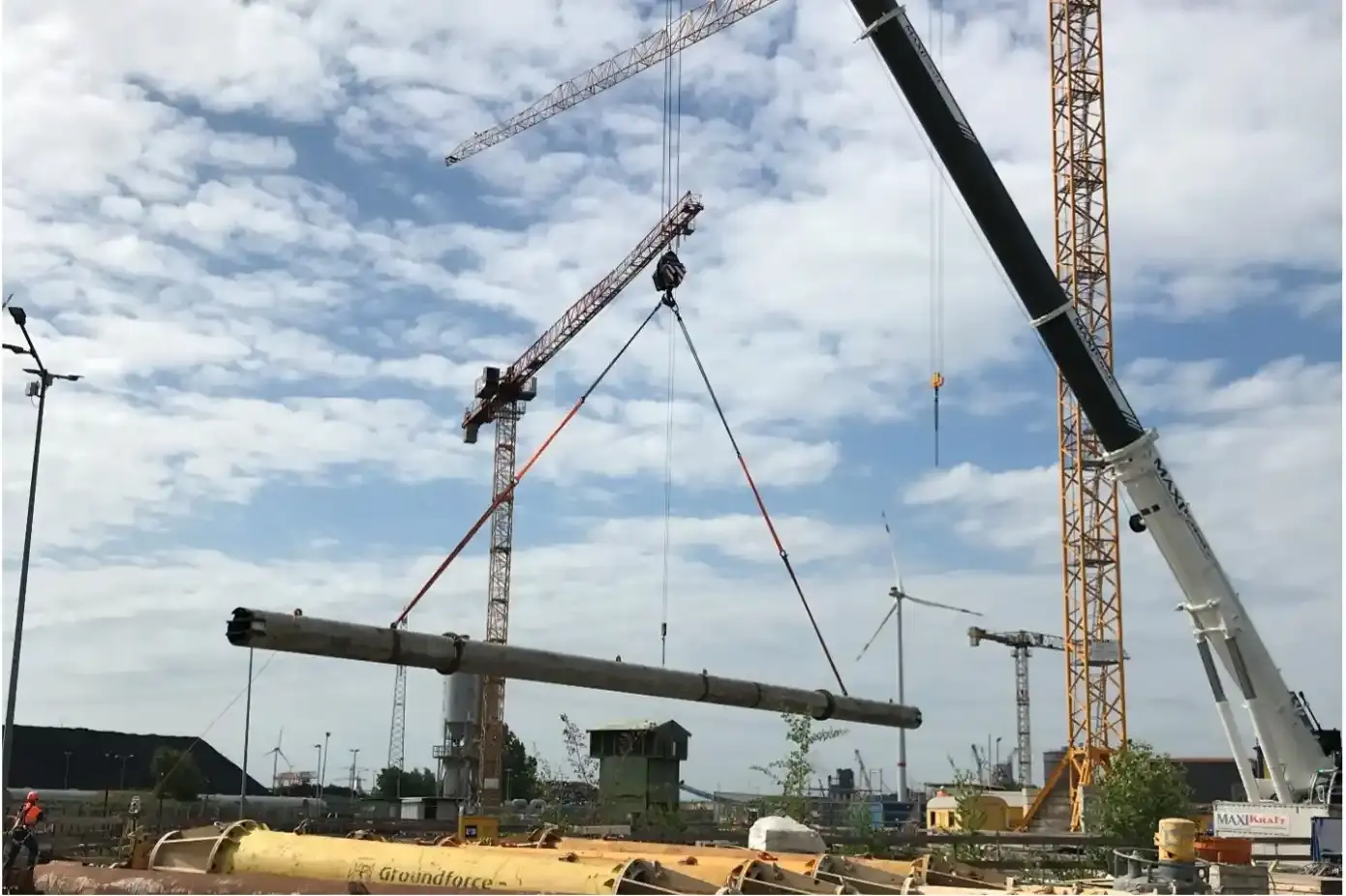Foto eines Krans, der auf einer großen Baustelle vor einem leicht bedeckten blauen Himmel ein langes Rohr in die Luft hebt.