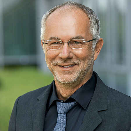 Portrait eines Mannes mit kurzem grauen Haar, Dreitagebart und Brille im dunklen Anzug mit Krawatte vor einem unscharfen Hintergrund.