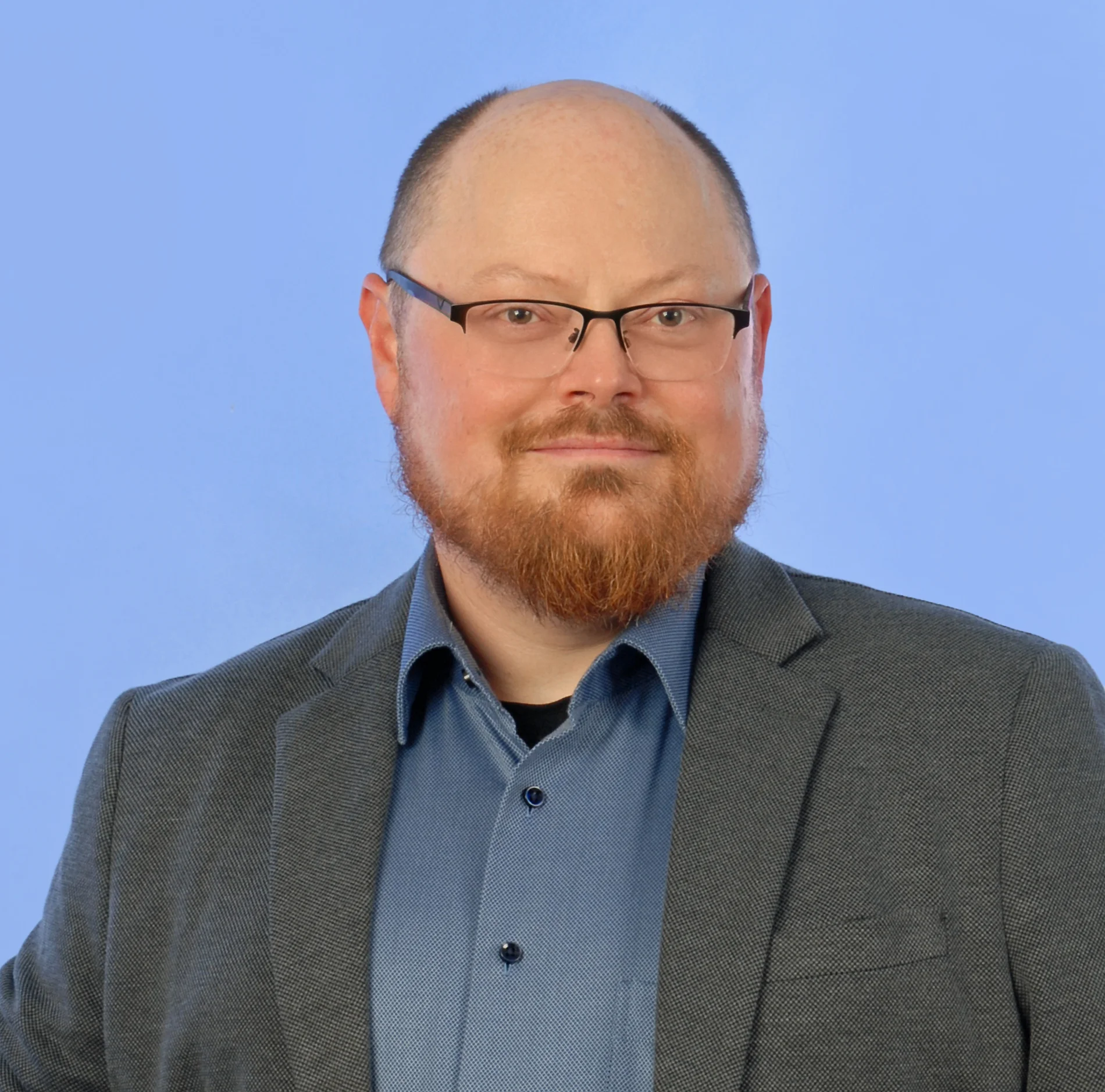 Mann mit Brille, kurzen Haaren und Bart lächelt in die Kamera und trägt ein dunkles Sakko über einem dunkelblauen Hemd. Der Hintergrund ist blau.