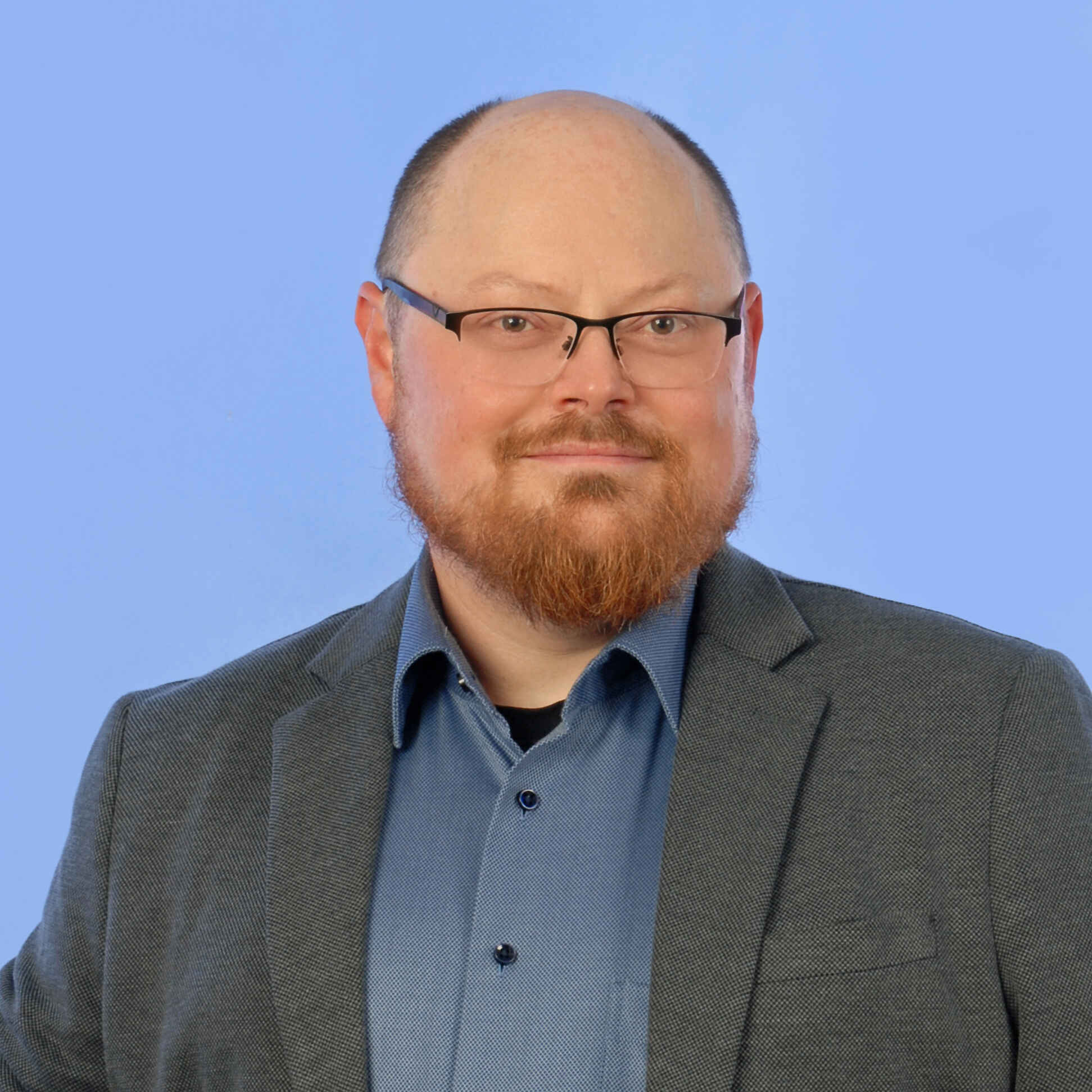 Mann mit Brille, kurzen Haaren und Bart lächelt in die Kamera und trägt ein dunkles Sakko über einem dunkelblauen Hemd. Der Hintergrund ist blau.