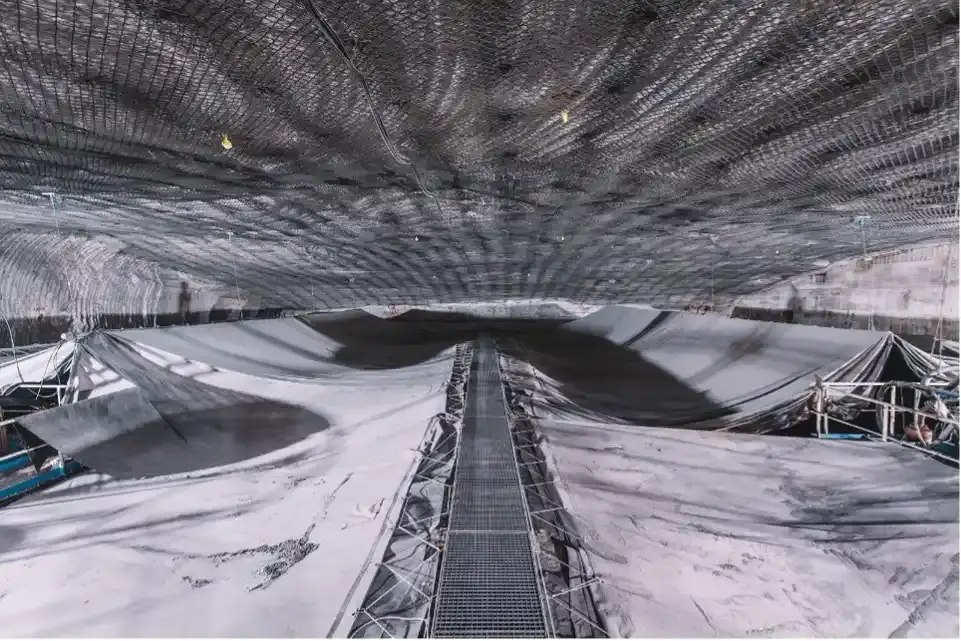 Das Bild einer Kammer in einem Salzbergwerk. Mittig verläuft ein Steg aus Metallgittern. Links und rechts des Stegs sind weißgraue Planen gespannt, die jeweils wie einer Art Zeltdach die darunterliegenden Becken bedecken. An der Abbaudecke aus Salzgestein sind Gitternetze zum Schutz vor herabfallenden Gesteinsbrocken verbaut.