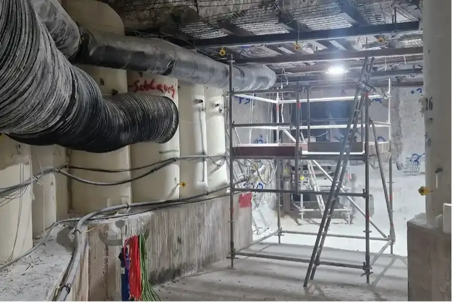 Das Bild zeigt den derzeitigen Arbeitsbereich. Auf der linken und rechten Seite befinden sich Stützbauwerke aus Salzbeton. Hinter einem Gerüst ist im hinteren Bereich die Fassungsstelle zu erkennen. An der Wand aus Salz ist eine schwarze Plane befestigt, um die Fassungsstelle zu schützen. Davor ist ein kleines Stehpodest aus Metall aufgestellt. Die gefasste Lösung wird über eine schwarze Rohrleitung geleitet. Das Rohr erinnert vom Durchmesser an einen größeren Gartenschlauch.