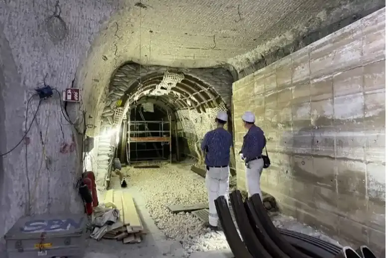Zwei Bergleute stehen in einer Strecke unter Tage mit dem Rücken zur Kamera. Sie blicken in einen Tunnel aus Metallstreben. In dem Tunnel ist ein Gerüst aufgebaut. Rechts neben den Bergleuten befindet sich eine Mauer aus Salzbeton. Hinter ihnen liegen gebogene Metallstreben auf dem Boden.