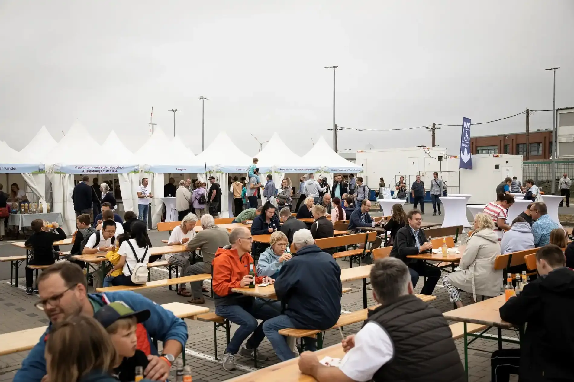 Menschen sitzen auf Holzbänken an Holztischen. Im Hintergrund stehen Zelte und Menschen an Stehtischen.