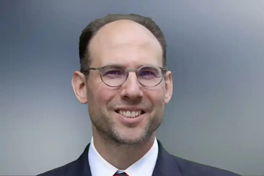 Ein Mann mit Brille, weißem Hemd und dunklem Sakko lächelt in die Kamera.