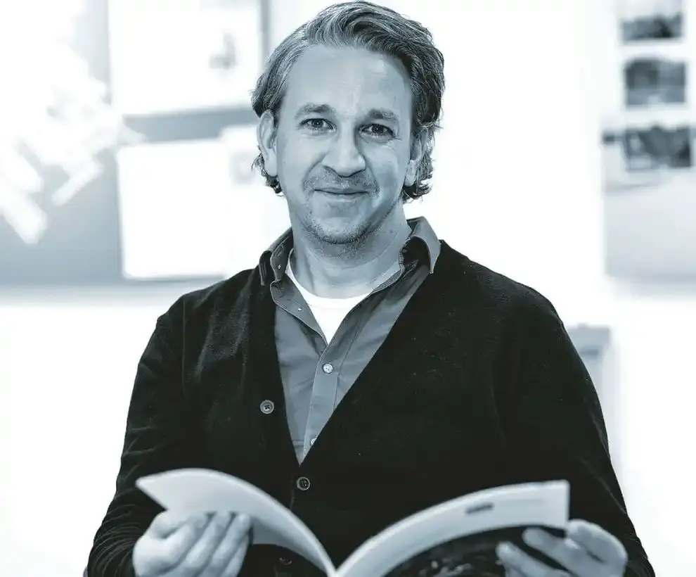 Schwarz-Weiß-Foto eines Mannes im Portrait. Dieser hat mittellange Haare, einen Dreitagebart und hält eine Broschüre in der Hand. Der Mann trägt ein Hemd unter einer zugeknöpften Jacke.