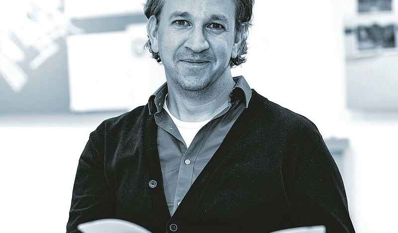 Schwarz-Weiß-Foto eines Mannes im Portrait. Dieser hat mittellange Haare, einen Dreitagebart und hält eine Broschüre in der Hand. Der Mann trägt ein Hemd unter einer zugeknöpften Jacke.