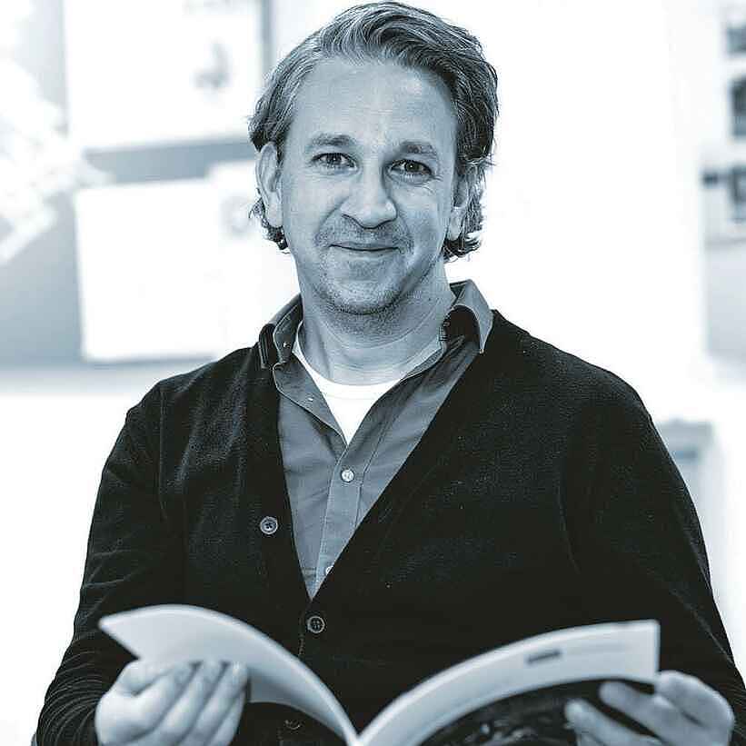 Schwarz-Weiß-Foto eines Mannes im Portrait. Dieser hat mittellange Haare, einen Dreitagebart und hält eine Broschüre in der Hand. Der Mann trägt ein Hemd unter einer zugeknöpften Jacke.