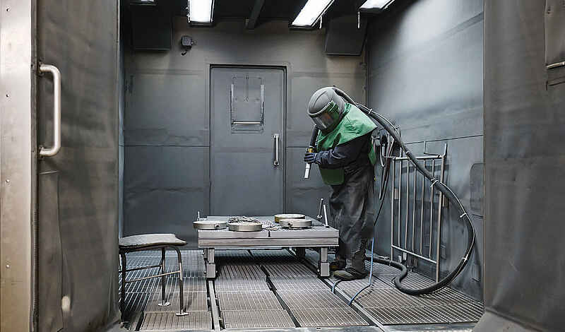 Person in Schutzanzug und Helm arbeitet mit Sandstrahlgerät in einer geschlossenen Metallkabine.