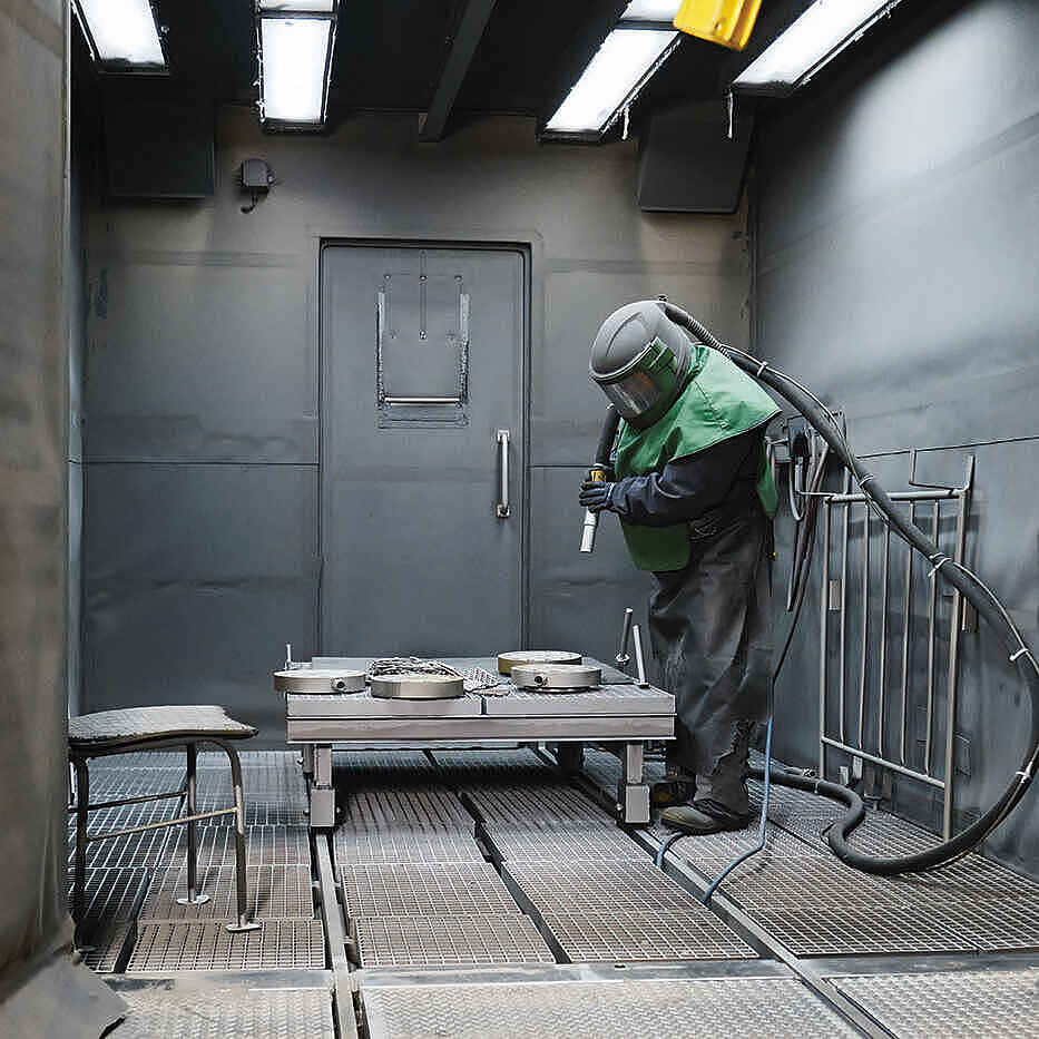 Person in Schutzanzug und Helm arbeitet mit Sandstrahlgerät in einer geschlossenen Metallkabine.