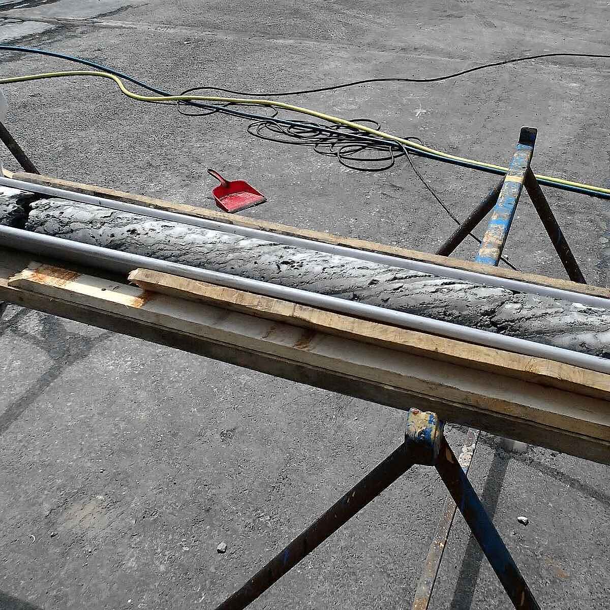 Ein zylindrischer Gesteinskörper liegt in einer Metallrinne auf zwei Böcken.