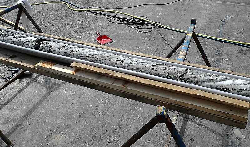 Ein zylindrischer Gesteinskörper liegt in einer Metallrinne auf zwei Böcken.