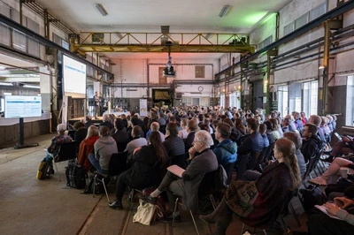 Auf dem Foto sind zahlreiche sitzende Menschen verschiedenen Alters zu sehen, die in einer Halle zur Bühne blicken. 