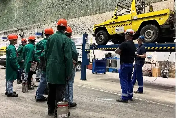Eine Gruppe von Menschen in grünen Kitteln steht vor zwei Männern in blauer Arbeitskleidung. Im Hintergrund steht ein gelbes Auto auf einer Hebebühne.