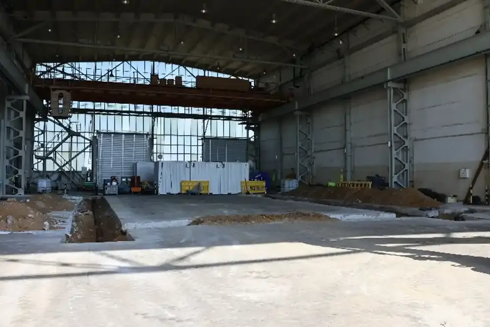 Der Innenbereich einer großen Industriehalle. Der Betonboden ist an einigen Stellen offen, sodass die dort verlegten Rohre zu erkennen sind.
