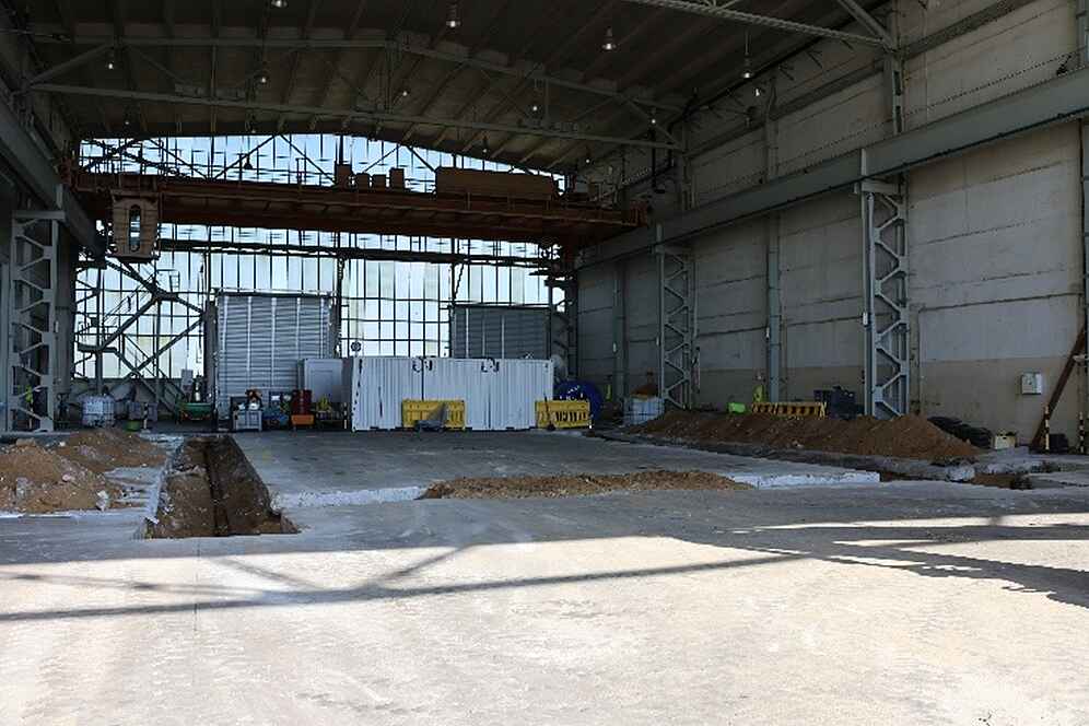 Der Innenbereich einer großen Industriehalle. Der Betonboden ist an einigen Stellen offen, sodass die dort verlegten Rohre zu erkennen sind.