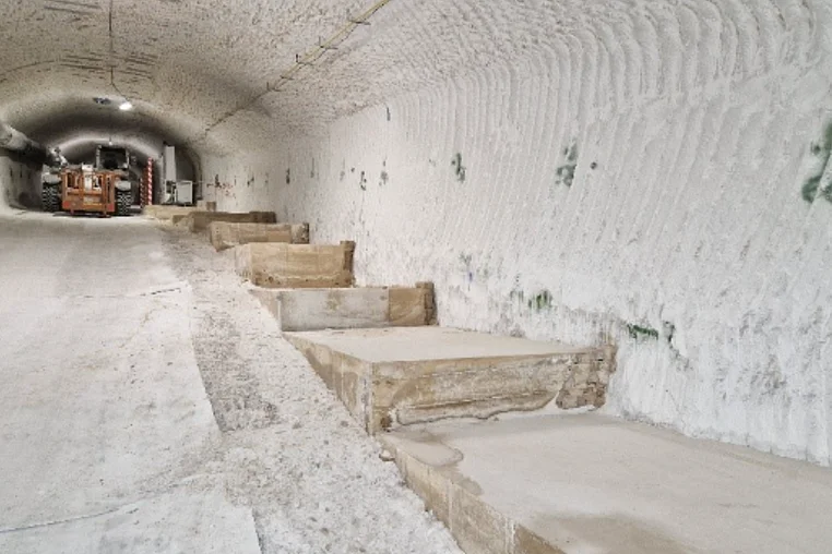 Das Bild zeigt eine ansteigende Strecke im Salzbergwerk. Auf der linken Bildseite befindet sich eine ebene Fahrbahn. Im oberen Bereich der Fahrbahn steht ein gelbes Fahrzeug. Im rechten Streckenbereich befinden sich mehrere, treppenartig angelegte Fundamente aus Beton. Die weißen Decken- und Wandbereiche weisen typische wellenartige Frässpuren auf. 