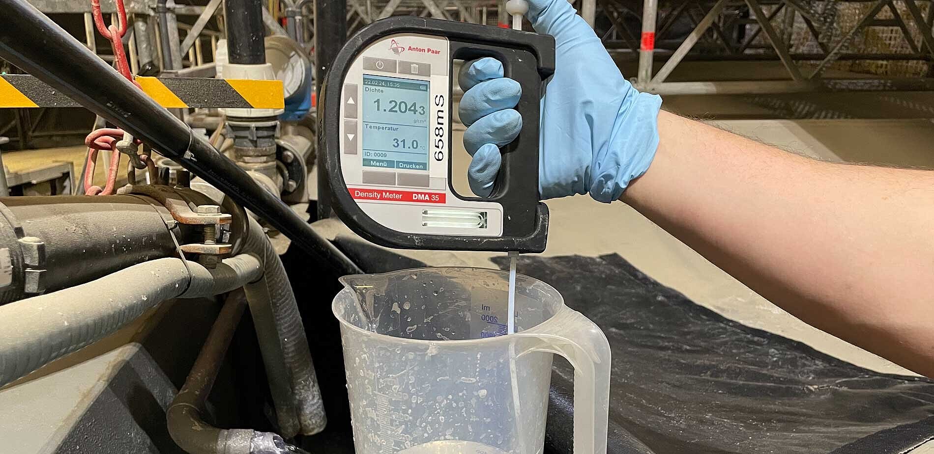 A gloved hand holds a measuring device in a vessel containing a clear liquid.