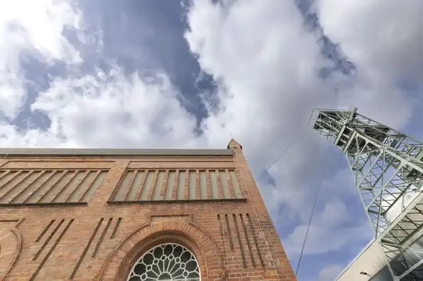 Das Foto zeigt ein rotes Backsteingebäude sowie das Stahlgerüst eines Förderturms vor einem blauen Himmel mit weißen Wolken.