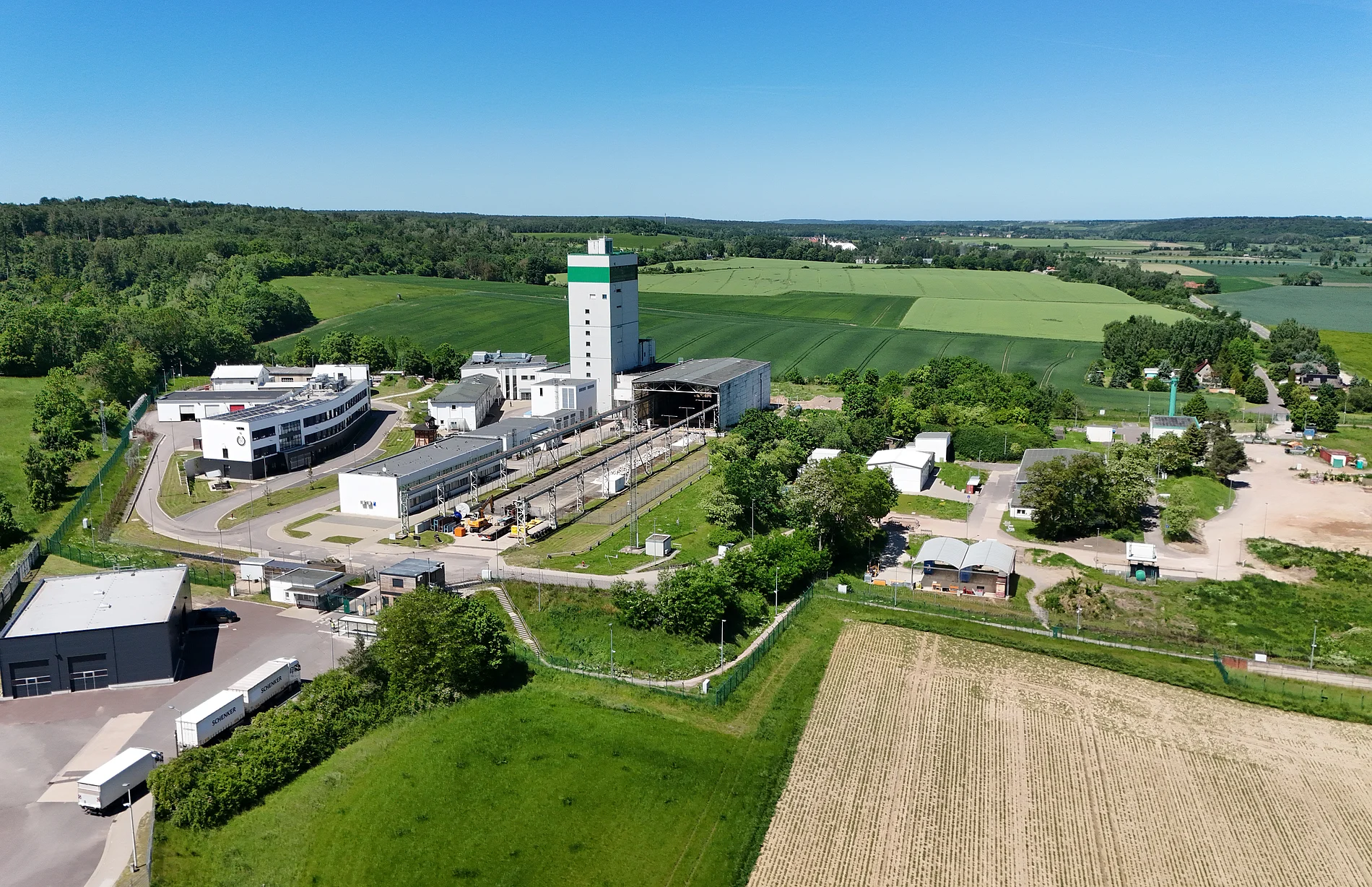 Das Endlager Morsleben fotografiert aus der Luft. Das Betriebsgelände samt weißem Förderturm ist umgeben von grünen Feldern und Wälder bei blauem Himmel.