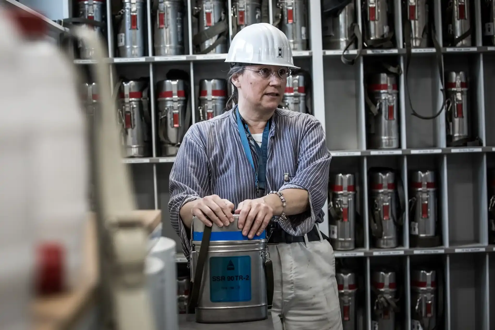 Eine Frau mit weißem Schutzhelm steht vor einem Regal mit silberfarbenen Sauerstoffselbstrettern. Ein Exemplar steht vor ihr auf einem Tisch. Diese Geräte versorgen Personen unter Tage beispielsweise im Brandfall mit Sauerstoff.