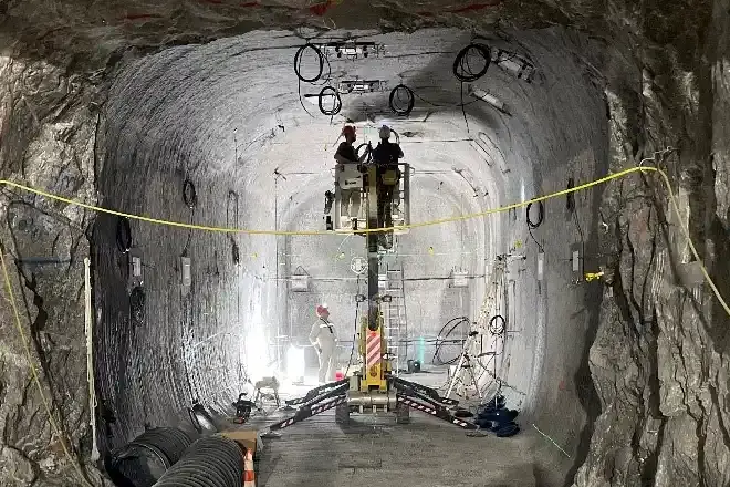 Foto einer Strecke unter Tage in einem Salzbergwerk. Zwei Personen befinden sich im Korb eines Krans und arbeiten an der Decke. Überfall verlaufen Kabel.