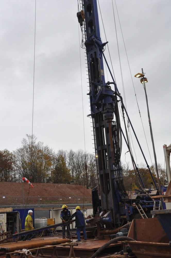 Foto einer mobilen Bohranlage vor einem Gebiet mit Bäumen. Mehrere Personen mit Schutzhelmen befinden sich in unmittelbarer Nähe zur Bohranlage.