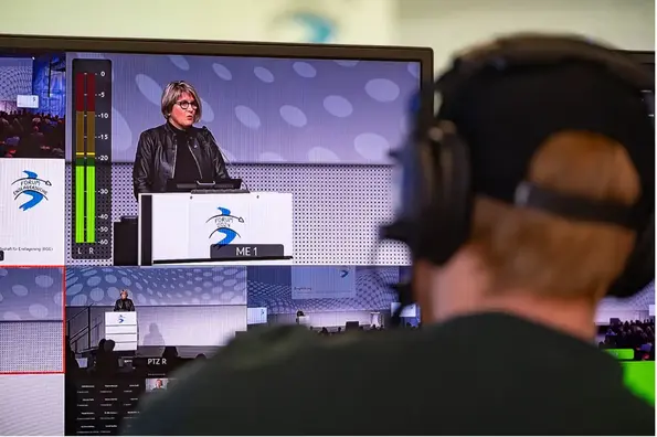 Ein Techniker blickt auf einen Bildschirm, auf dem eine Frau in ein Mikrofon spricht. 