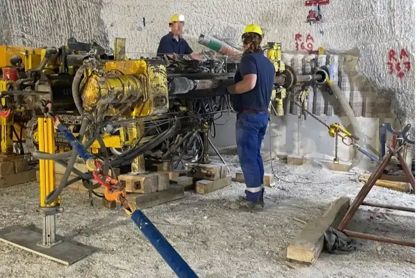 Zwei Personen in blauen Arbeitsanzügen und gelben Sicherheitshelmen arbeiten unter Tage in einem Salzbergwerk an einer gelbgrauen Maschine mit vielen Schläuchen. Die Maschine steht auf Holzblöcken.