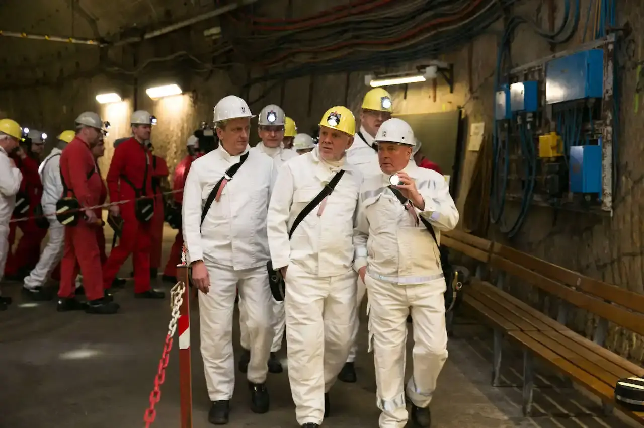 Gruppe von Personen in Schutzkleidung und Helmen bei einer Untertage-Begehung, im Hintergrund weitere Personen in roten Overalls.