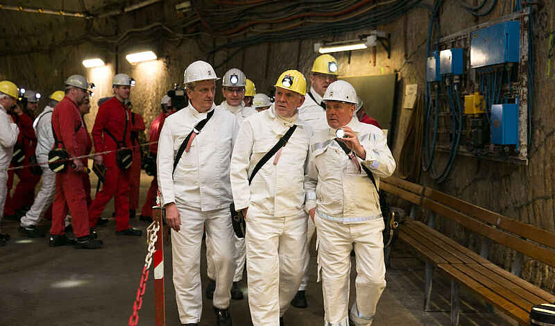 Gruppe von Personen in Schutzkleidung und Helmen bei einer Untertage-Begehung, im Hintergrund weitere Personen in roten Overalls.
