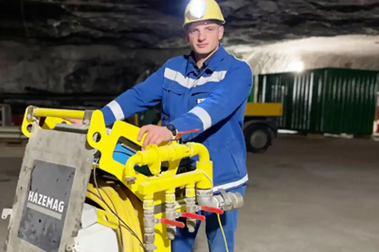 Ein junger Mann in blauer Bergmannskleidung und gelbem Schutzhelm blickt in die Kamera. Er befindet sich unter Tage in einem Bergwerk und seine Hände ruhen auf einer gelben Apparatur mit mehreren Anschlüssen für Schläuche.