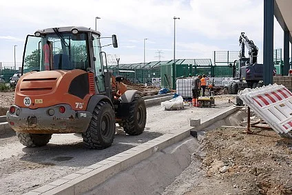 Ein Radlader steht auf dem Unterbau einer Zufahrtsstraße, davor führen Bauleute Straßenbauarbeiten durch. 
