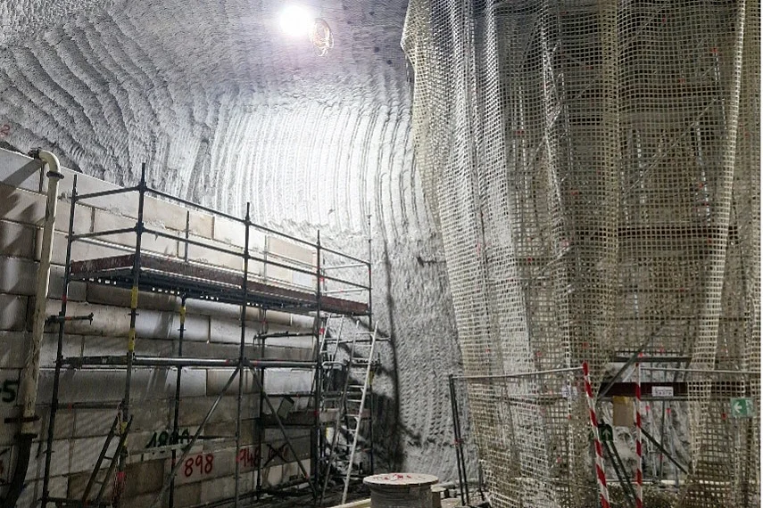 Das Bild zeigt einen rund 15 Meter hohen Raum im Bergwerk. Der Raum wird auf der linken Seite auf halber Höhe durch eine Mauer aus Beton-Blocksteinen abgegrenzt. Vor der Mauer ist ein Gerüst aufgebaut. Über die Mauer verläuft eine Rohrleitung in den dahinterliegenden Bereich. Auf der echten Bildseite ragt ein mit einem Netz abgehängtes Gerüst bis über die Decke hinaus in einen offenen Schacht hinein.