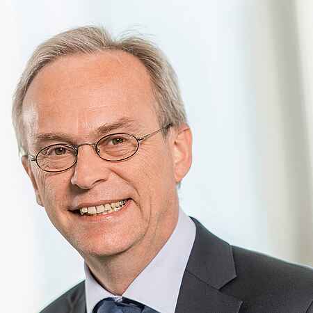 Portrait eines Mannes mit grauen kurzen Haaren und Brille. Er trägt ein hellblaues Hemd, ein schwarzes Sakko und eine Krawatte.