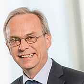 Portrait eines Mannes mit grauen kurzen Haaren und Brille. Er trägt ein hellblaues Hemd, ein schwarzes Sakko und eine Krawatte.