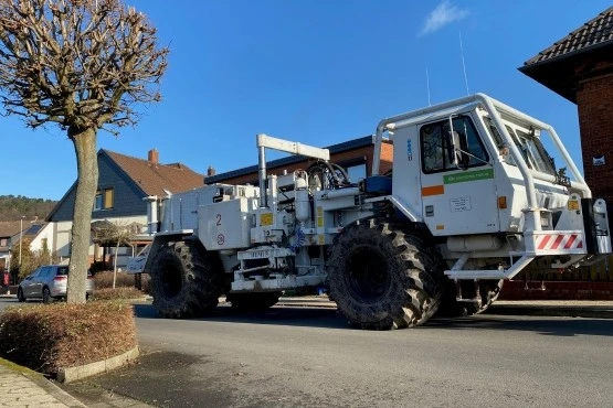Das Foto zeigt eine schmale Straße in einem Dorf. Die Sonne scheint und der Himmel ist blau. Ein großer weißer LKW steht auf der Straße. Dessen Reifen sind ähnlich groß wie die an der Hinterachse eines Traktors.