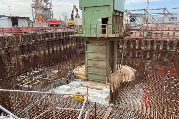 In einer großen rechteckigen Vertiefung im Boden - ähnlich einer Baugrube - sind zahlreiche Metallstreben sowie nachträglich ins Bild eingefügte rote Markierungen zu sehen. In der Mitte der Vertiefung ragt ein grüner Turm in die Höhe. Im Hintergrund sind ein Bagger und Baugerüste zu sehen. 