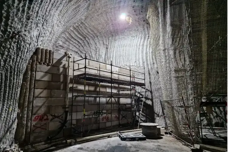 Das Bild zeigt einen großen offenen Bereich im Bergwerk. Die Decke und die Wände sind aus Salzgestein und weisen wellenartige Frässpuren auf. Aus der Bildmitte nach links erstreckt sich über die halbe Höhe des offenen Bereiches eine große Wand aus aufeinander gebauten Blocksteinen aus (Sorel-) Beton. Die Mauer grenzt einen dahinterliegenden Hohlraum ab. Vor der Mauer steht ein Gerüst. Aus der Bildmitte nach rechts erstreckt sich eine Art Vorhang aus einem Netz. Hinter dem Netz steht ein Gerüst, das bis unter die Decke reicht.